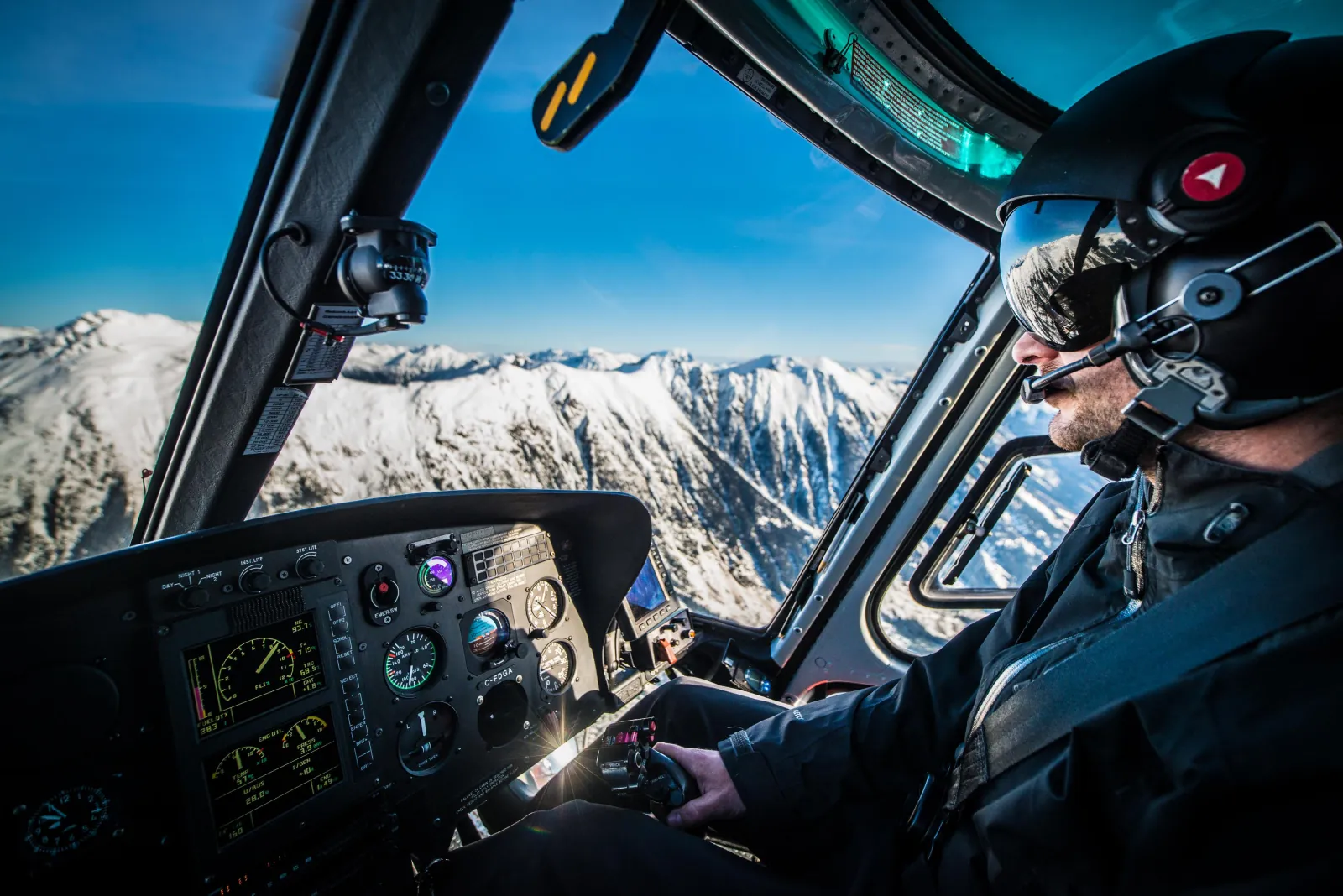 Helicopter pilot in a helmet at the controls, with snowy mountain ridges through the windscreen
