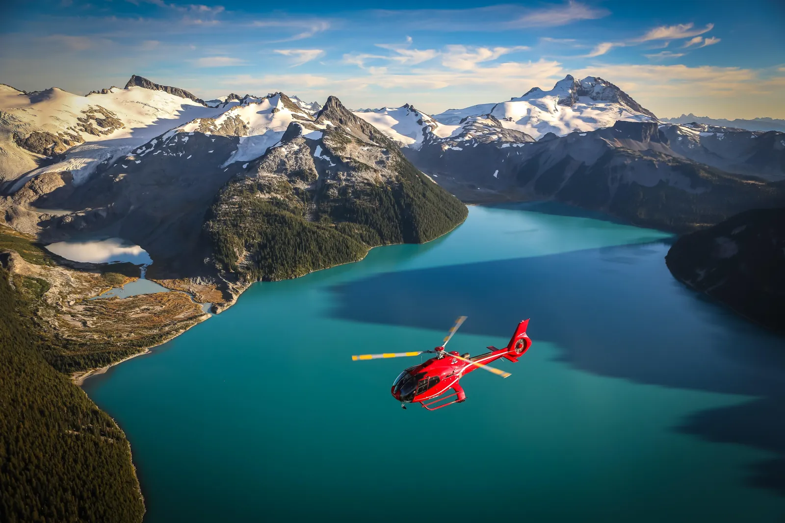 Red helicopter flying over turquoise Garibaldi Lake below snowy glaciated peaks