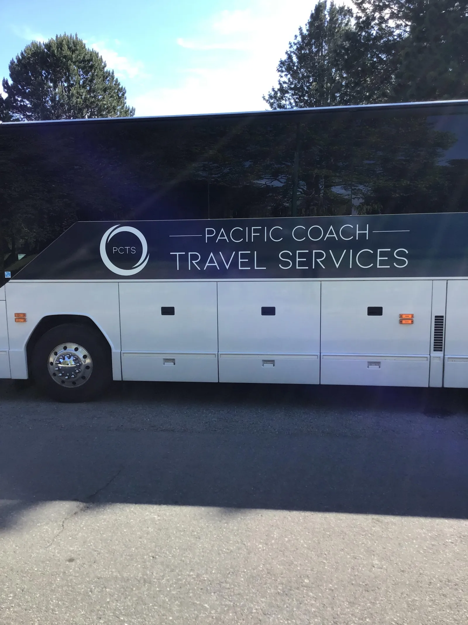 Side of a white Pacific Coach Travel Services coach parked in the sun