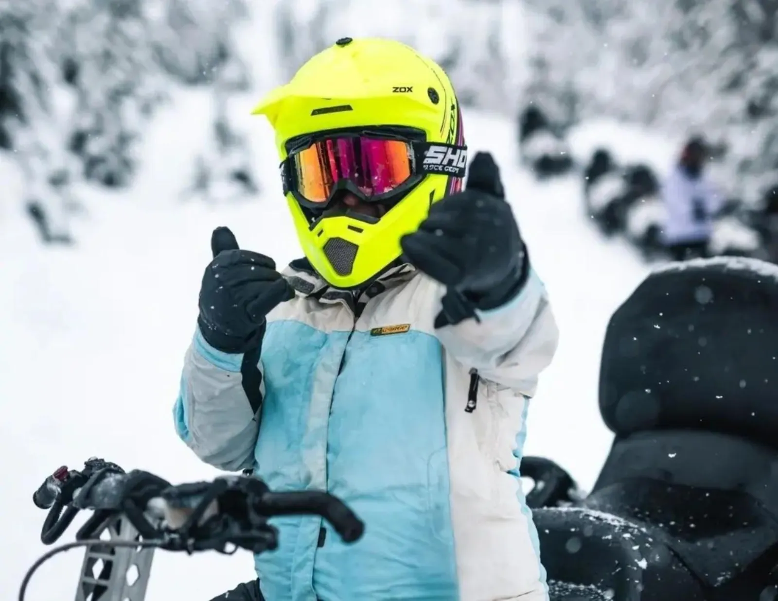 Young rider in a yellow helmet and goggles giving a thumbs up beside a parked snowmobile