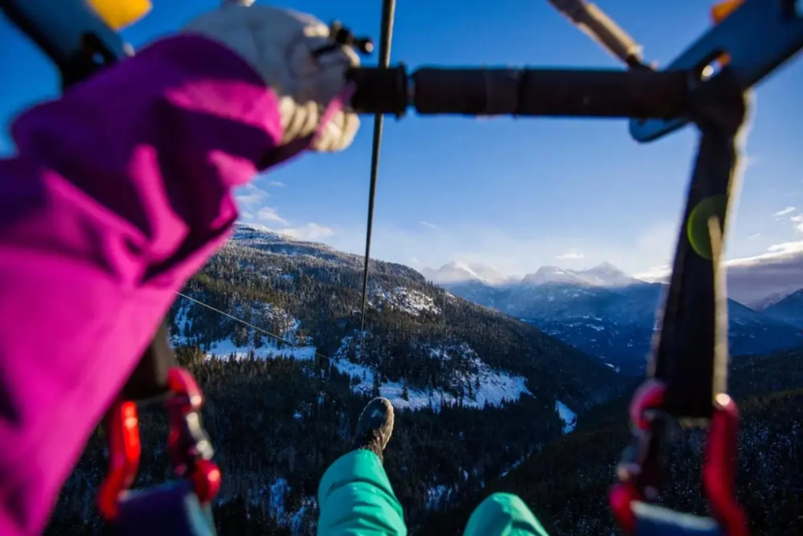 Rider's-eye view from a seated zipline harness over snowy forest toward mountains