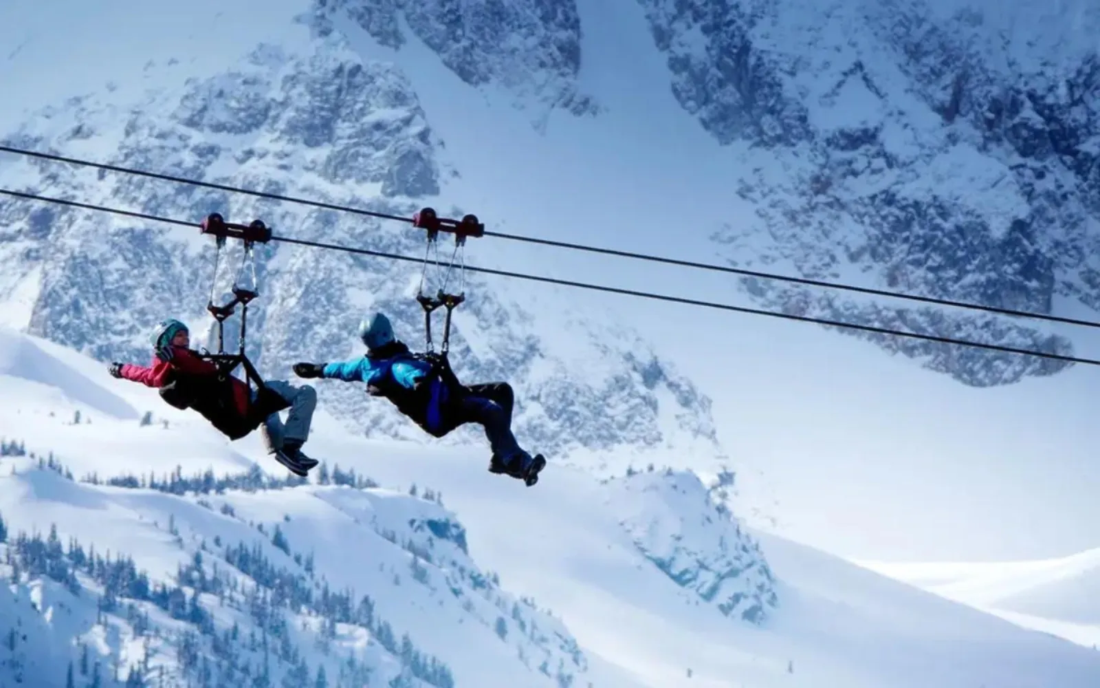 Two riders in seated harnesses on tandem lines in front of a snowy mountain face in winter