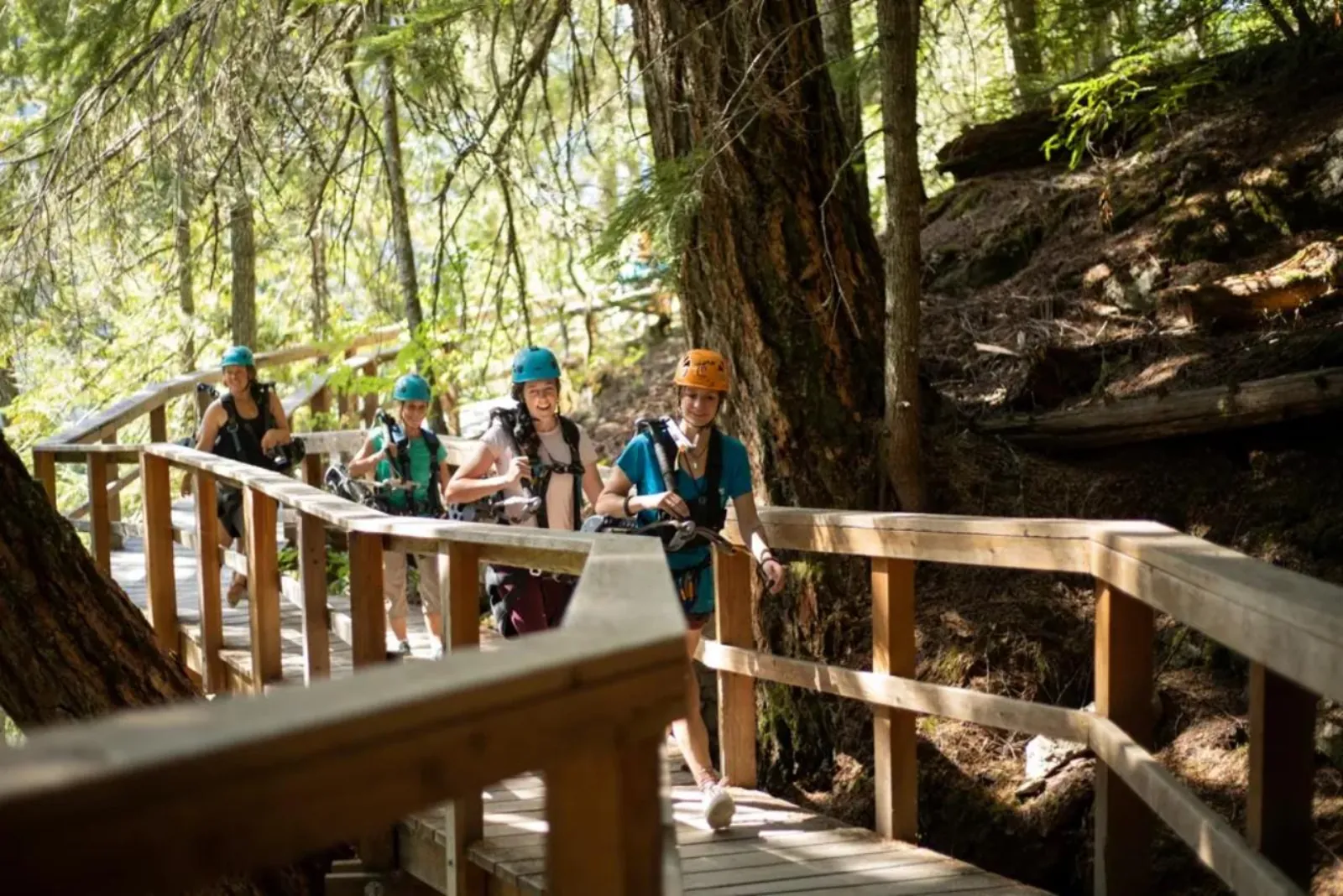 Four riders in helmets carrying zipline trolleys along a wooden boardwalk in the forest