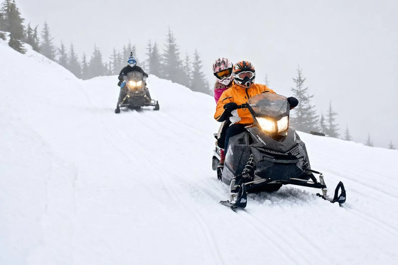 Driver and passenger on a snowmobile descending a snowy trail in mist, a second rider behind
