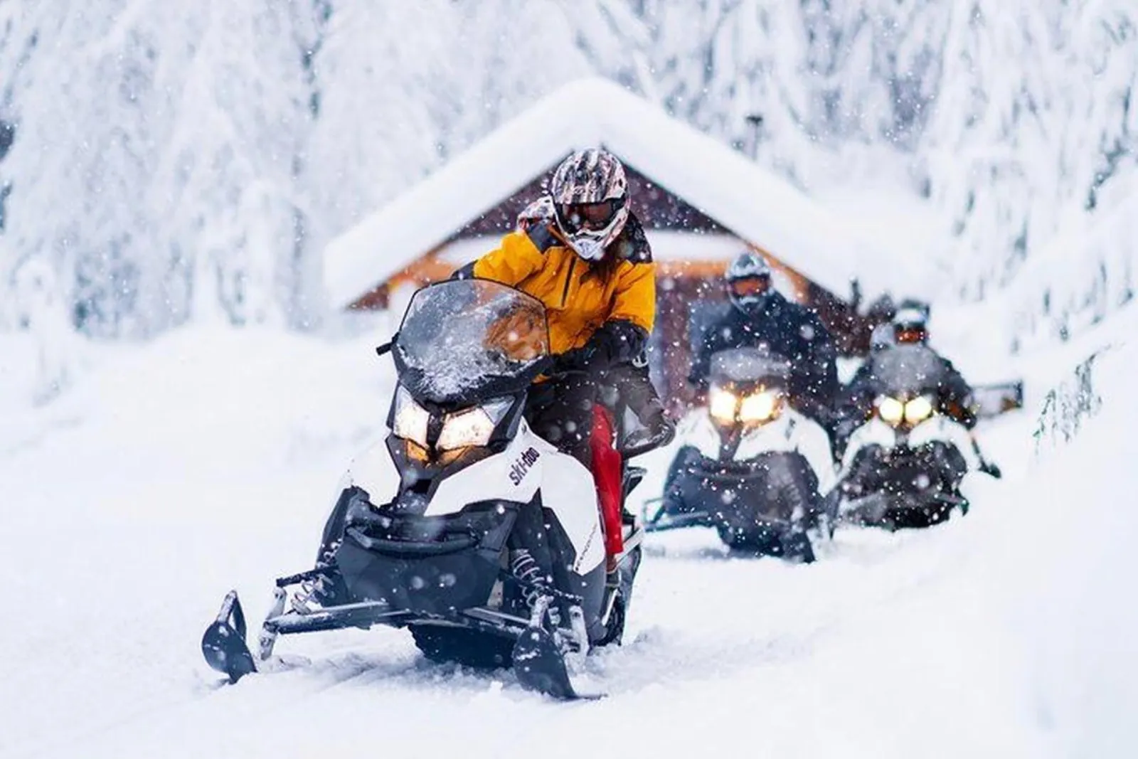 Riders on white Ski-Doo snowmobiles leaving a snow-covered log cabin in falling snow
