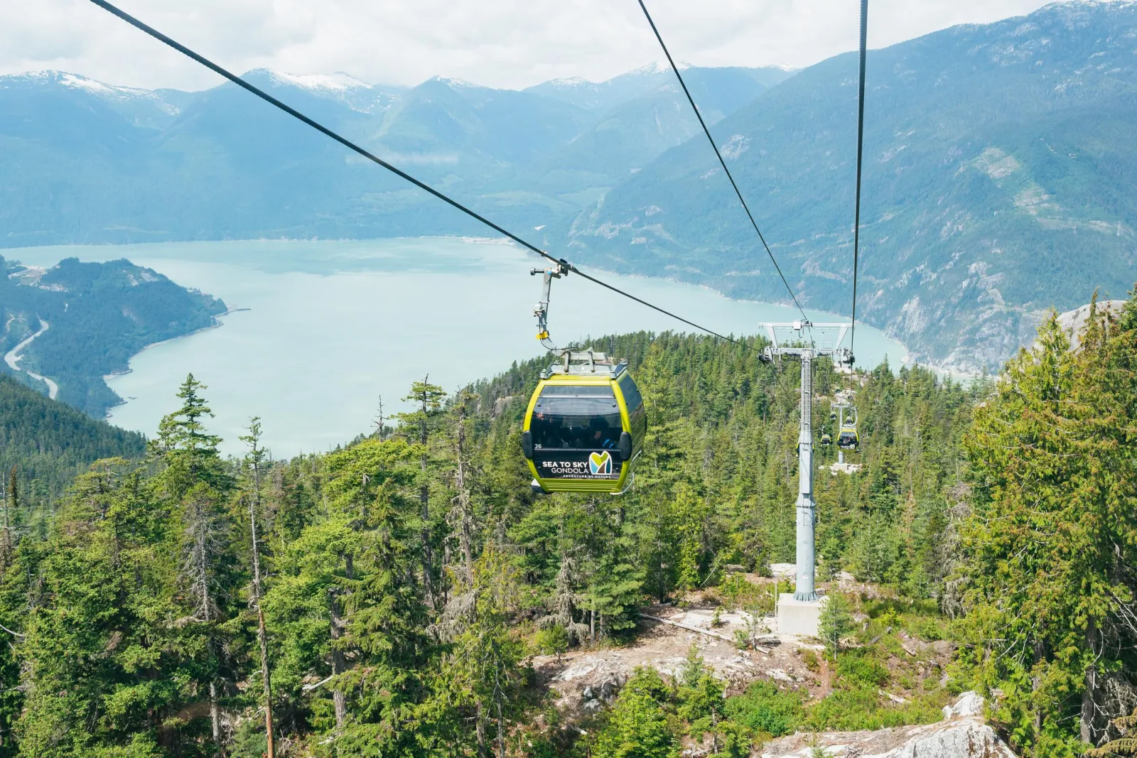 Yellow Sea to Sky Gondola cabin rising over forest with Howe Sound and snowy peaks behind