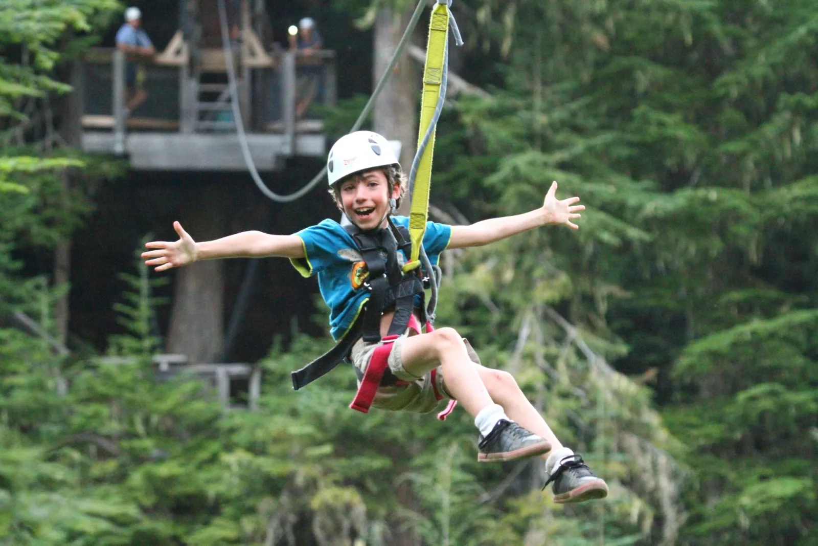 Smiling boy with arms outstretched on a zipline, a wooden platform in the trees behind