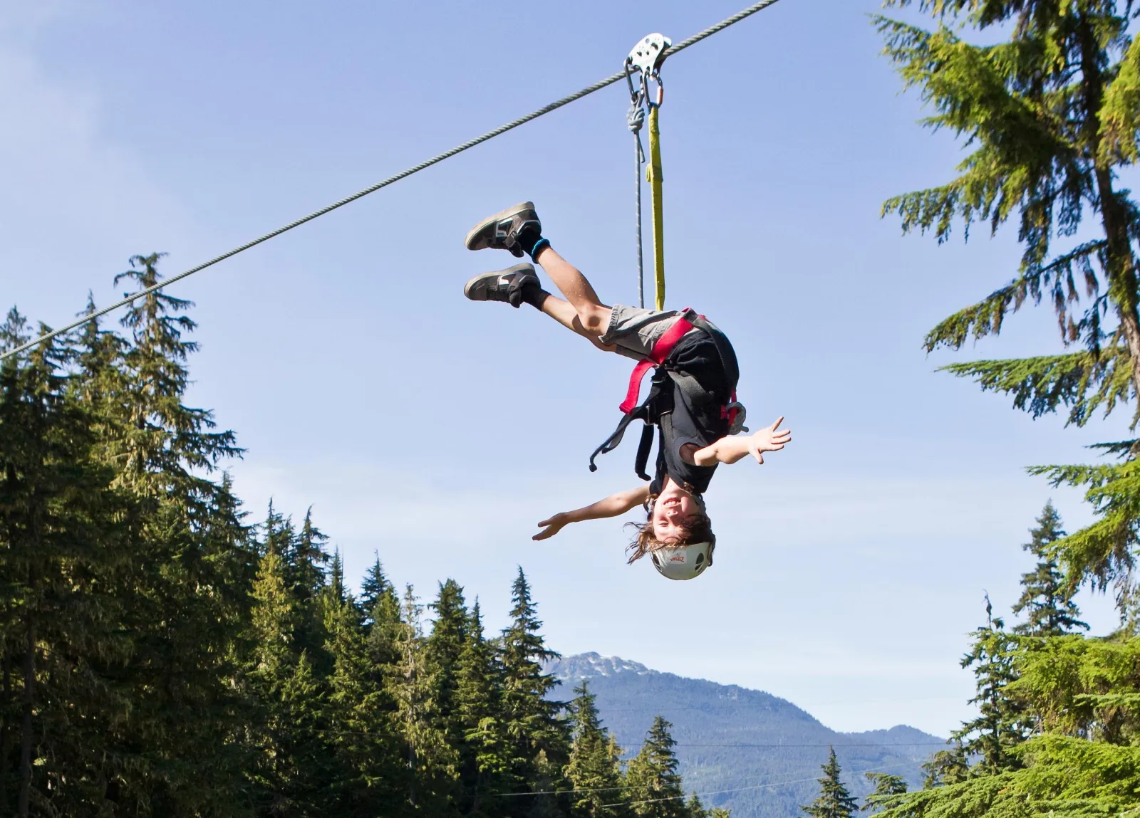 Child hanging upside down on a zipline against blue sky, forest and a mountain behind
