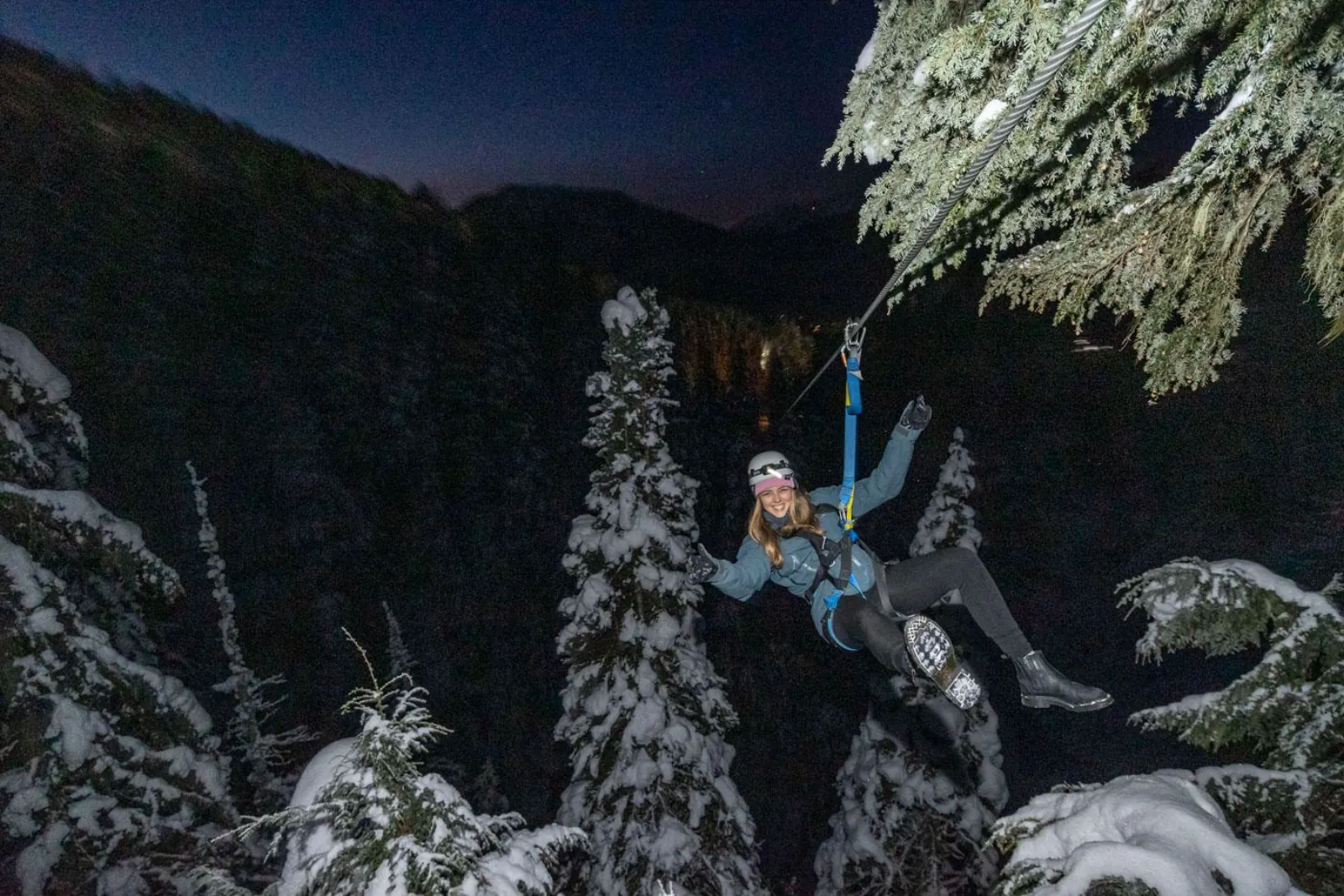 Smiling rider on a zipline at night between snow-covered conifers