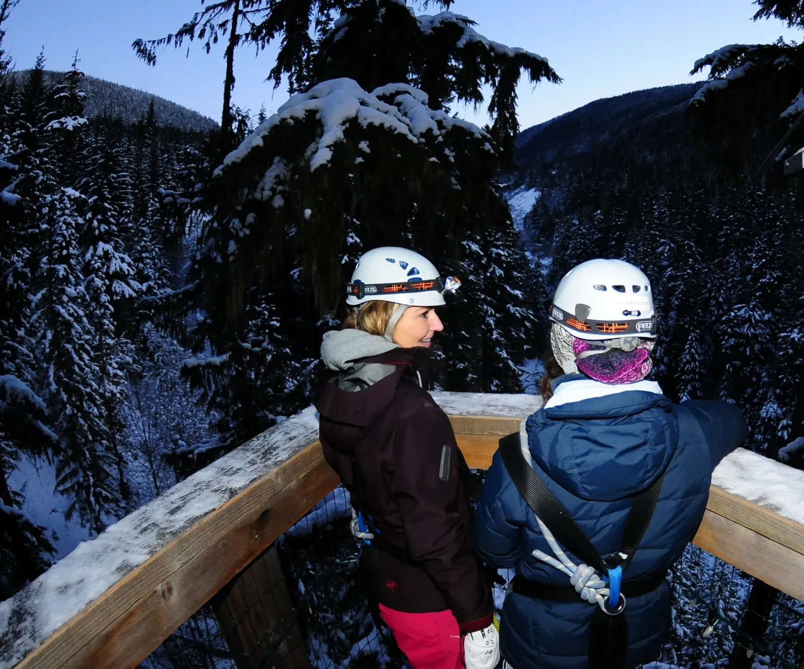 Two riders with headlamps on a snowy treetop platform looking over a forested valley at dusk