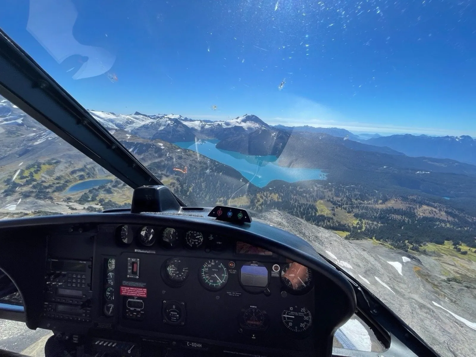 View through a helicopter windscreen over alpine meadows toward a turquoise lake and glaciated peaks