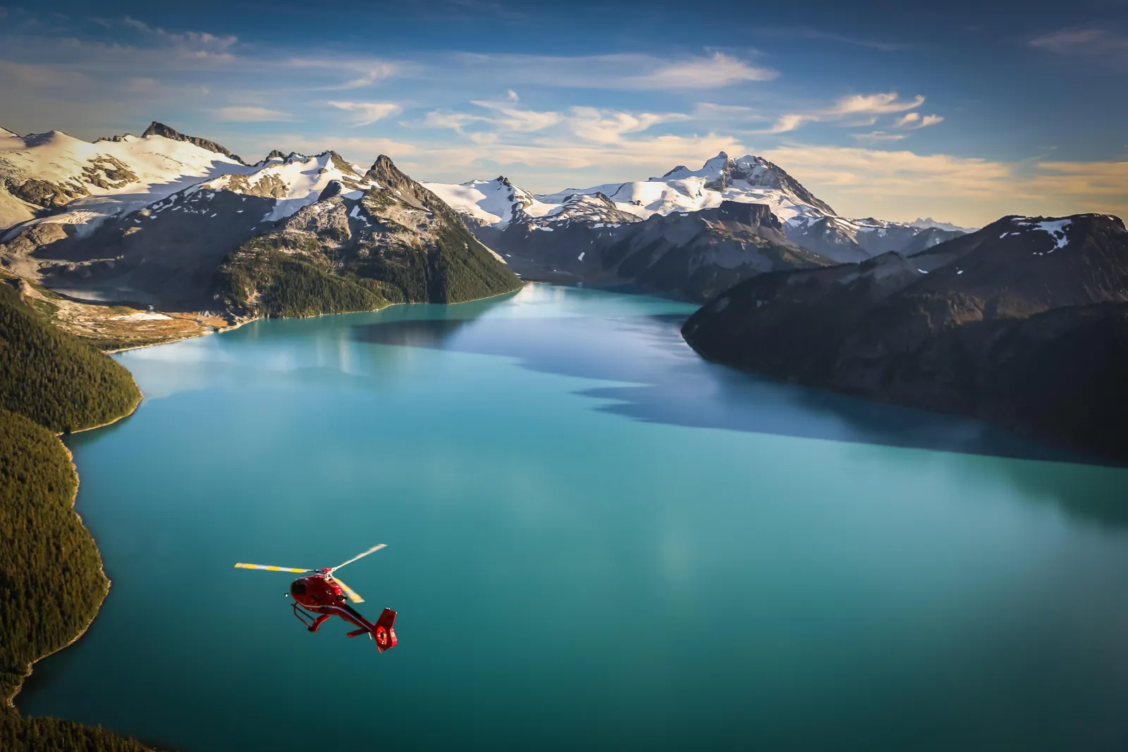 Red helicopter over a turquoise alpine lake ringed by snowy peaks and forest