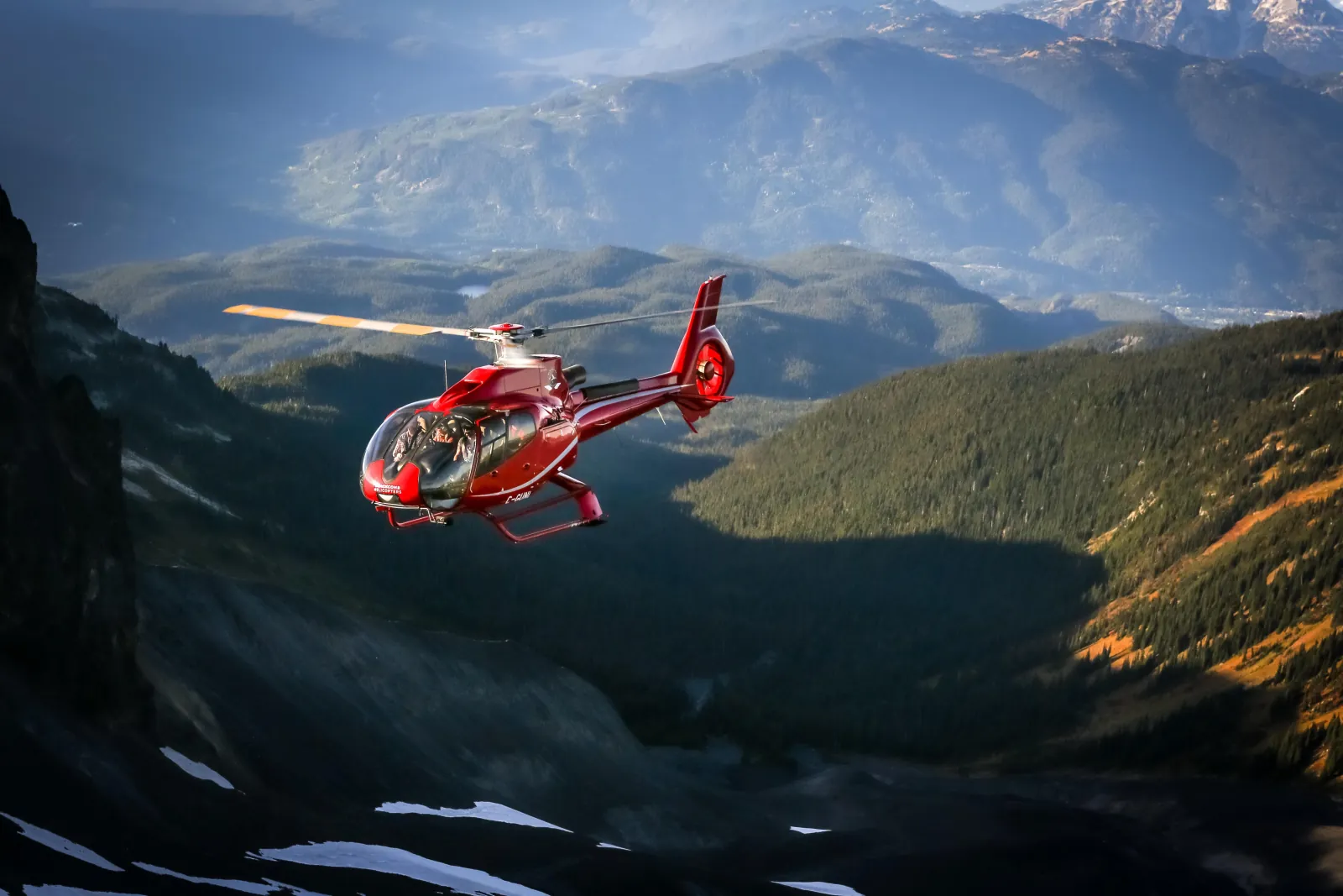 Red helicopter with passengers aboard flying over a forested valley in autumn light