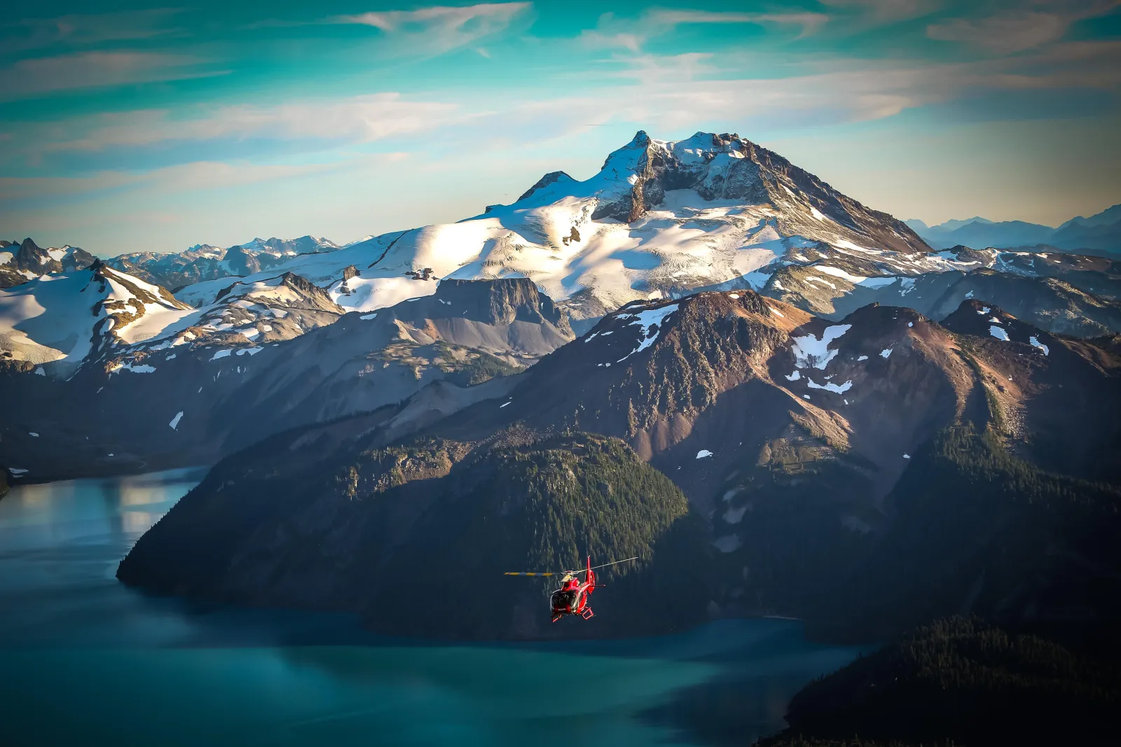 Small red helicopter over a blue lake beneath a broad glaciated volcanic peak
