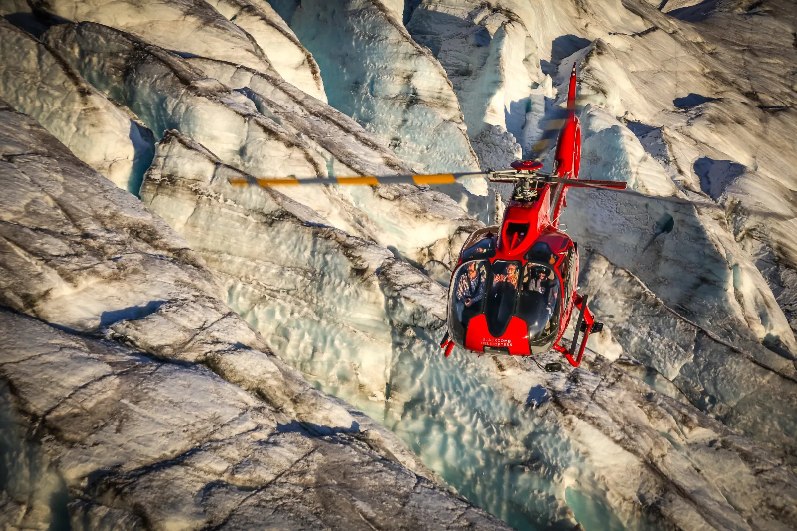 Passengers seen through the bubble windscreen of a red helicopter flying low over glacier ice