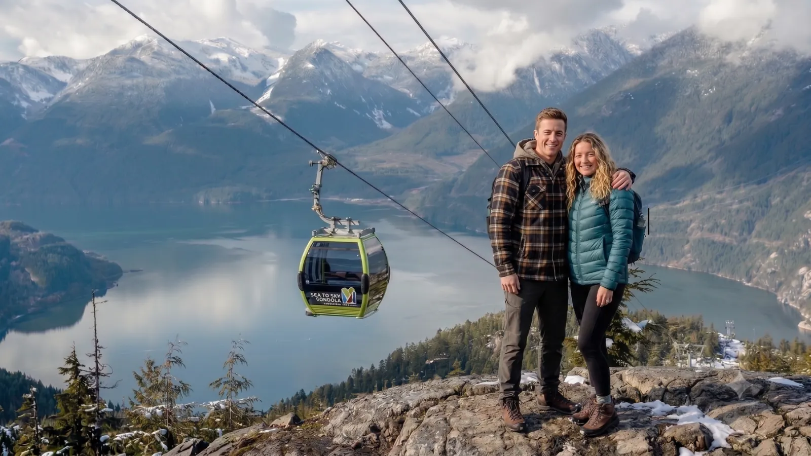 Couple on a rocky summit viewpoint with a Sea to Sky Gondola cabin and Howe Sound behind