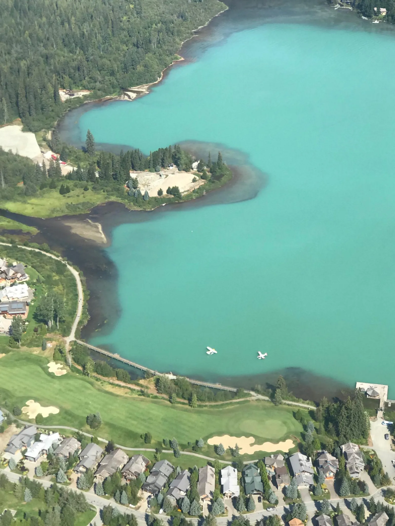 Two seaplanes on turquoise Green Lake beside a golf course and Whistler homes, from the air