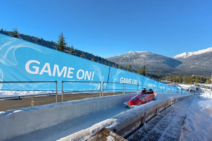 Red bobsled slowing on the sunlit finish stretch with snowy mountains behind