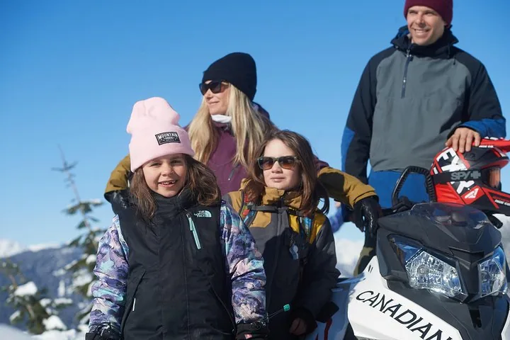 Family of four in winter clothing beside a Canadian Wilderness snowmobile with mountains behind
