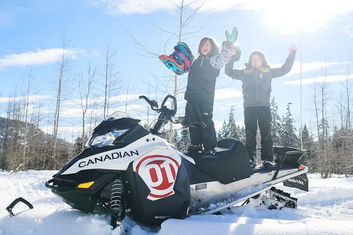 Two children cheering while standing on a parked snowmobile in a snowy clearing