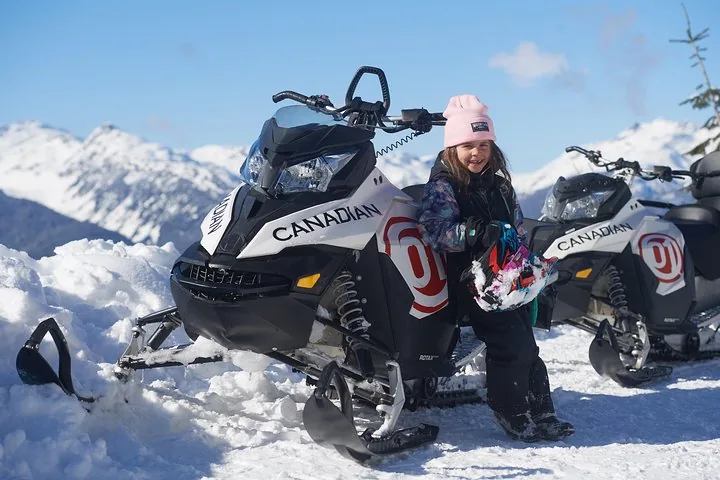 Young girl in a pink hat leaning on a parked snowmobile with snowy peaks in the distance