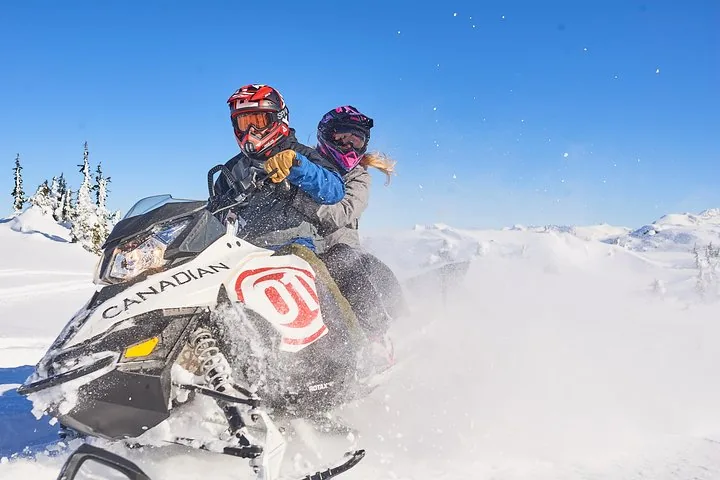 Driver and passenger in helmets and goggles on a white Canadian snowmobile under a blue sky