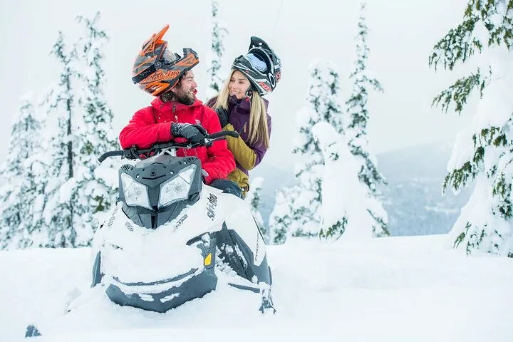 Couple in helmets sitting on a parked snowmobile among snow-covered trees