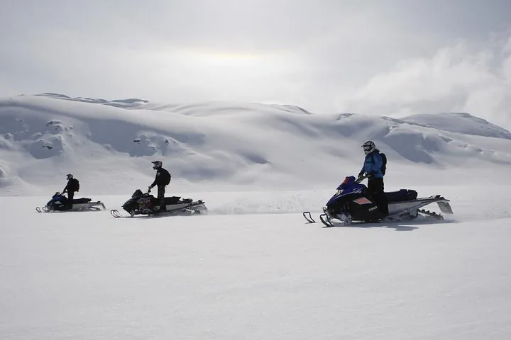 Three riders standing on their snowmobiles on a wide snowfield under grey skies