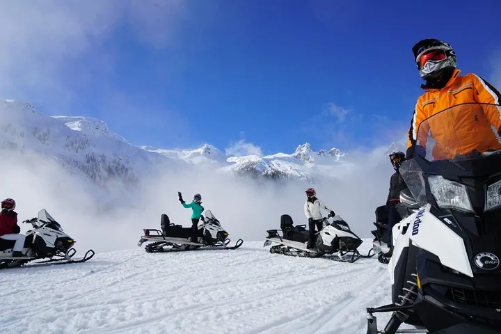 Group of riders stopped at a snowy lookout above low cloud with white peaks behind