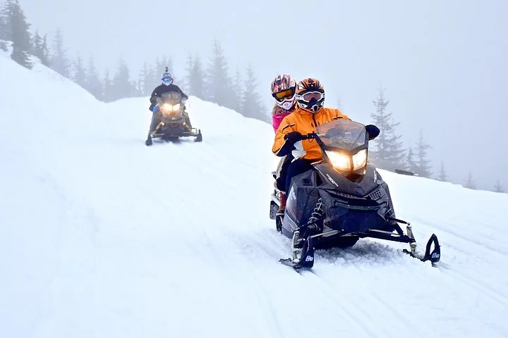 Driver and passenger in helmets on a snowmobile climbing a misty trail, another rider behind