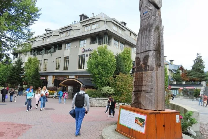Visitors walking through Whistler Village past a carved wooden pole on a cloudy summer day